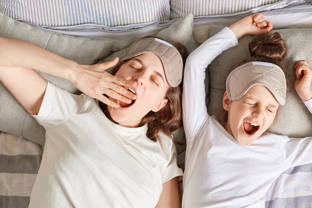 Sleepy mother and daughter sleeping together waking up wearing sleep masks and white shirts yawning lazy morning enjoying happy time together. - Foto, imagen