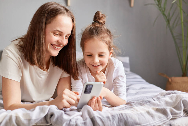 Smiling mother and adorable daughter using phone, relaxing on bed at home, little girl child with mum looking at smartphone screen with happy expression. - Photo, Image