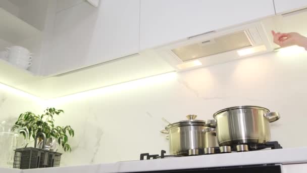 Een vrouw zet de motorkap boven het fornuis aan om te koken. Nieuwe moderne keuken in witte kleuren. - Video