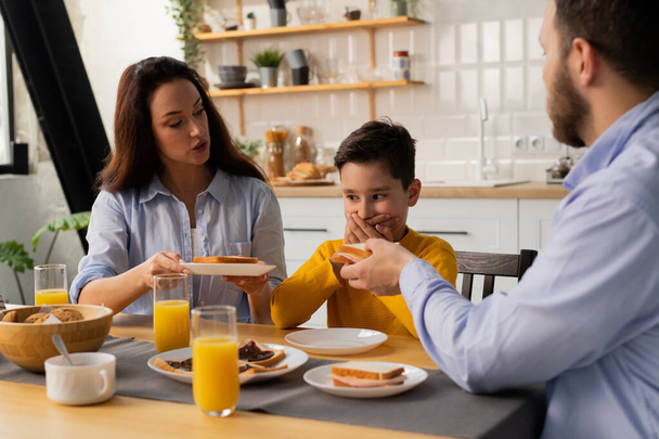 El niño se sienta a la mesa, cubriéndose la boca con las manos, y sus padres le dan sándwiches. Los padres están tratando de averiguar por qué su hijo se niega a comer. Foto de alta calidad - Foto, Imagen