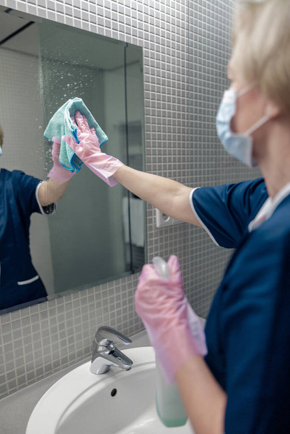 Cleaning lady cleans mirror in bathroom spraying detergent on surface. Housekeeping concept - Photo, Image
