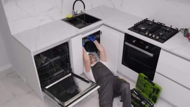 Fiatal vízvezeték-szerelő egyenruhában mosogatót javít a konyhában. Egy hívás egy vízvezeték-szerelő házába. 4k videó - Felvétel, videó