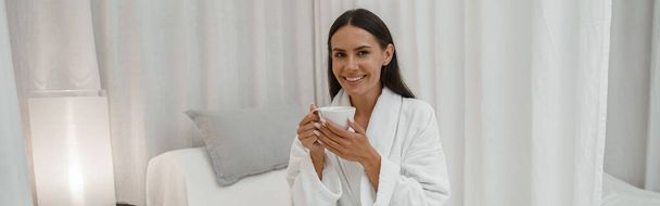 Szczęśliwa ładna kobieta w białym szlafroku korzystających podczas odpoczynku z filiżanką kawy w salonie spa - Zdjęcie, obraz
