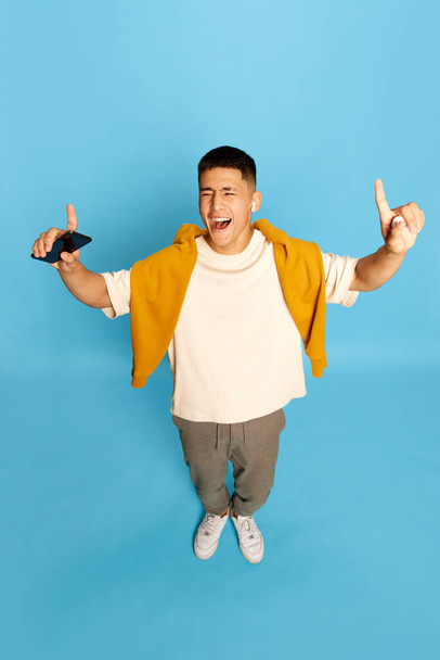 Portrait with excited young man, guy, student in stylish casual clothes listening to music via earpods and smiling over blue background. Top view. Concept of hobby, music, fashion, emotions, ad - Photo, Image