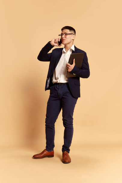 Business meeting. Portrait with one charming young man, office worker in fashion style costume, suit talking via cell phone over beige background. Career, human emotions, online, sales concept - Foto, Imagem