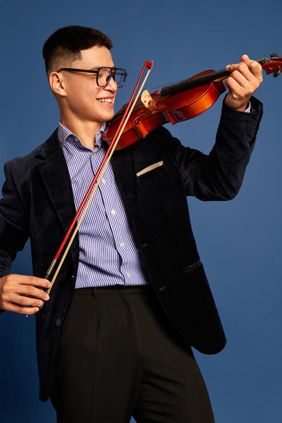 Portrait with professional, young musicians, violinist wearing costume holding fiddlestick and playing on violin and smiling over blue background. Concept of music, art, hobby, human emotions, ad - Fotó, kép