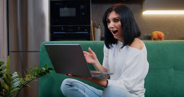 Happy and excited young caucasian woman winner looks at laptop celebrates online success sits on green sofa at home in modern kitchen background. - 写真・画像