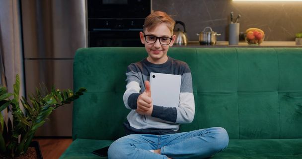 Portrait of 9 year old cute boy holding tablet device sitting on comfortable sofa in living room and gesturing thumbs up into camera - Photo, Image