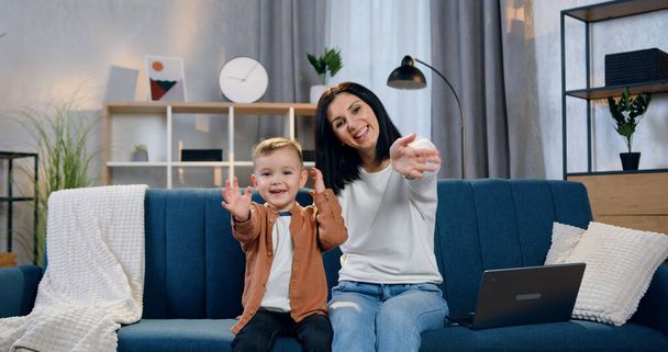 Adorable smiling happy mother and her little in high spirits son sitting together on the sofa at home and waving hands into camera to say hello - Photo, Image