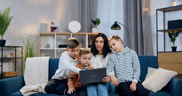 Attractive smiling carefree young mother explaining how to use laptop her three interested different ages sons when sitting with them on soft couch at home - 写真・画像