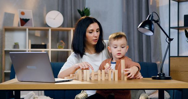 Portrait of pleasant happy loving mother which sitting at the table together with her small son and composing wooden blocks - Photo, Image