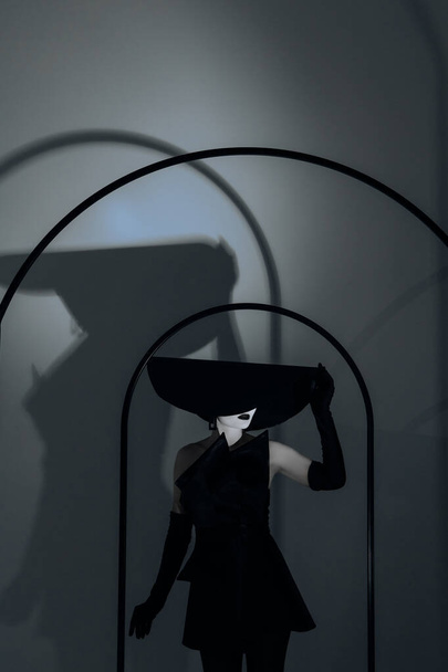 Giovane donna con il trucco sul viso posa in studio in elegante cappello a tesa larga, abito nero e guanti contro la sua ombra sul muro. - Foto, immagini