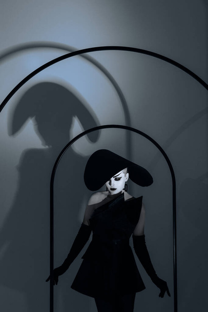 Fiatal nő smink az arcán pózol a stúdióban elegáns széles karimájú kalap, fekete ruha és kesztyűt neki árnyék a falon. - Fotó, kép