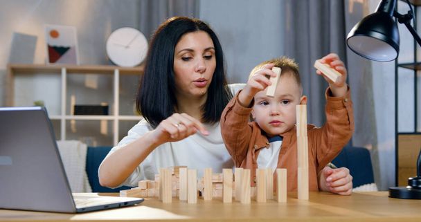 Lovely small curious boy sitting on his mothers knees and trying with her to compose a construction with wooden bricks on the table at home - Photo, Image