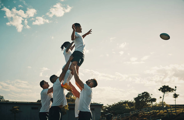 Rugby, team jump and lift for ball and teamwork, sport and training for game, fitness and exercise or sports performance with blue sky. Group, men and lifting man to catch and professional match. - 写真・画像