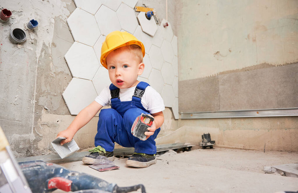 Adorable baby boy construction worker crouching down and looking at camera while holding tape measure. Cute kid wearing safety helmet and work overalls while working on home renovation. - Foto, imagen