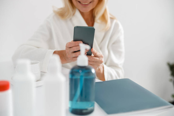 Unrecognizable blonde mature woman sitting in cosmetology center and uses cellphone surfing the net - Φωτογραφία, εικόνα