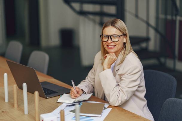 Woman accountant in eyeglasses making notes during work on laptop in cozy office. High quality photo - Foto, Bild