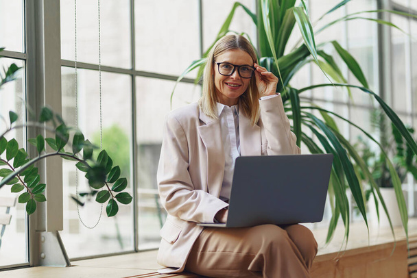 Smiling adult business woman working laptop while sitting in modern office. Blurred background - Фото, изображение