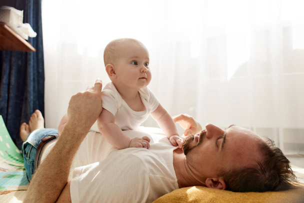 Счастливый отец держит своего новорожденного ребенка на руках, лежа на полу в яркой детской комнате. Отец смотрит на ребенка любящим взглядом. Счастливого детства и отцовства - Фото, изображение