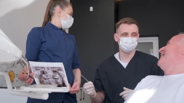 Dişçi koltuğunda oturan gri saçlı bir emekli diş sorunlarından bahsediyor. Diş muayenehanesindeki erkek dişçi dişlerin resmini tartışıyor. Sağlık, ilaç ve dişçilik - Video, Çekim