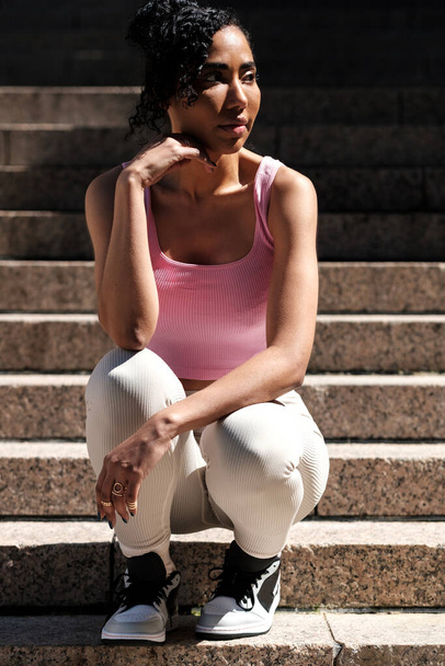 Attraktive Fitness-Frau posiert auf Treppen. Sie sitzt in gehockter Position und trägt rosa Sport-BH und Leggings. Sie sieht entspannt aus und schaut weg. - Foto, Bild