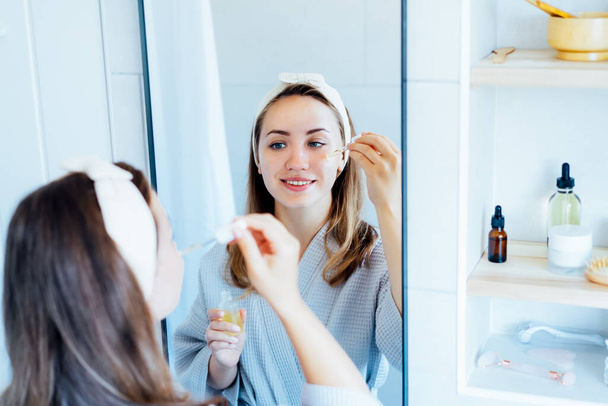 Young woman in bathrobe looking in the mirror, holding dropper with hyaluronic acid serum close to face and smiling. Skin hydrating. Cosmetic spa procedures. Beauty self-care at home. Selective focus - Photo, image