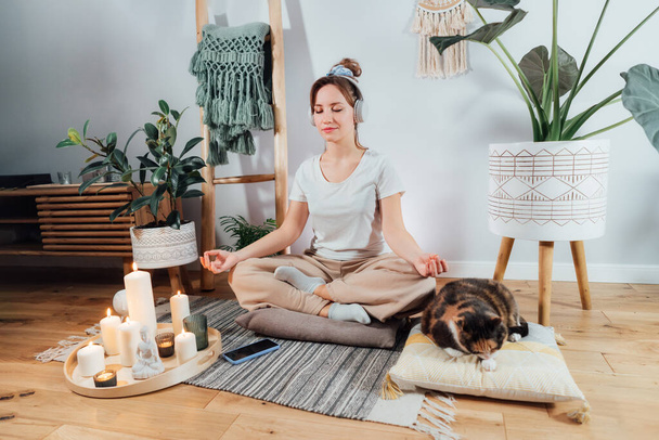 Young healthy woman practicing meditation at home, wearing headset, sitting in lotus position on floor cushion in modern Scandi interior home. Meditating, relaxed, eyes closed. Selective focus. - Foto, imagen