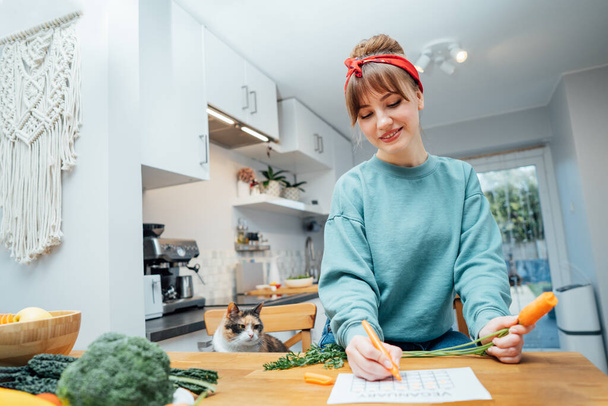 Young woman filling veganuary calendar while eating fresh carrot sitting on kitchen. Fresh vegetables and fruits on table. Vegan and vegetarian diet eating and cooking at home. Selective focus. - Фото, зображення