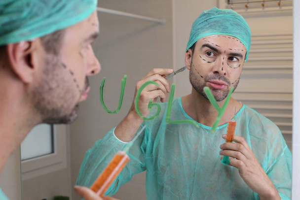 Porträt eines gutaussehenden jungen Mannes im chirurgischen Kostüm, der sich vor dem Spiegel auf eine Schönheitsoperation vorbereitet - Foto, Bild