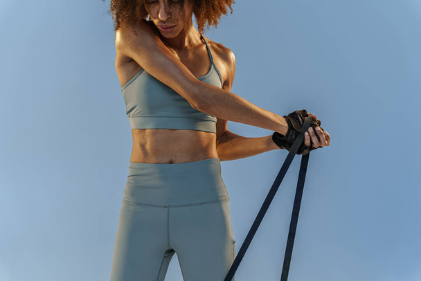 Strong woman in sportswear performs fitness exercises with resistance band on studio background - Photo, image