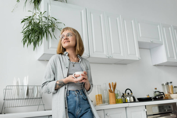 joyful young woman with short hair and bangs, eyeglasses and tattoo holding cup of morning coffee while looking away and standing in casual clothes next to dishes, kettle, kitchen appliances  - Fotografie, Obrázek