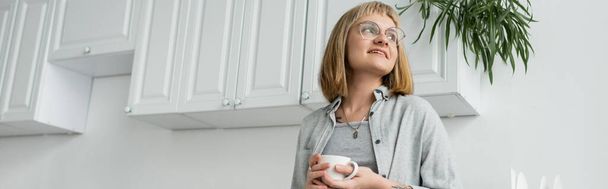 mujer joven positiva con pelo corto y flequillo, anteojos y tatuaje sosteniendo taza de café de la mañana mientras mira hacia otro lado y de pie en ropa casual al lado del gabinete de cocina blanco y la planta, pancarta  - Foto, Imagen