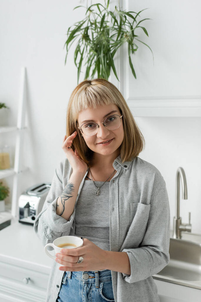happy young woman with bangs, eyeglasses and tattoo on hand adjusting short hair and holding cup of coffee while looking at camera, standing in casual clothes next to toaster, kitchen sink at home  - 写真・画像