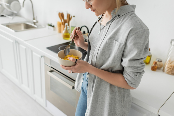 обрезанный вид молодой женщины в очках, держащей миску с кукурузными хлопьями и ложкой, стоя в повседневной серой одежде рядом с кухонной техникой в размытой белой кухне дома  - Фото, изображение