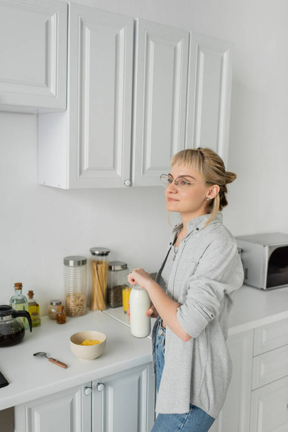 mujer joven en gafas graduadas y pelo corto con flequillo sosteniendo botella con leche cerca de tazón con copos de maíz mientras hace el desayuno y de pie en ropa casual al lado de electrodomésticos de cocina en casa  - Foto, imagen