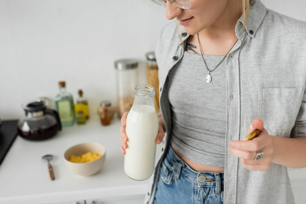 abgeschnittene Ansicht einer tätowierten Frau, die eine Flasche mit frischer Milch in der Nähe einer verschwommenen Schüssel mit Cornflakes und Löffel hält, während sie frühstückt und in Freizeitkleidung neben Küchengeräten zu Hause steht  - Foto, Bild