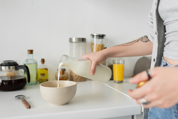 vista recortada de mujer joven tatuada sosteniendo botella mientras vierte leche fresca en un tazón con copos de maíz en la encimera de la cocina mientras hace el desayuno y de pie en ropa casual en la cocina moderna  - Foto, Imagen