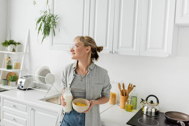 татуированная молодая женщина с челкой в очках держит бутылку со свежим молоком и миску с кукурузными хлопьями во время завтрака и глядя в сторону, стоя в повседневной одежде на современной кухне  - Фото, изображение