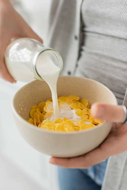 Genç bir kadının elinde şişeyle kaseye mısır gevreğiyle taze süt dökerken ve modern mutfakta günlük kıyafetlerle kahvaltı yaparken görüntüsünü kapatın.   - Fotoğraf, Görsel