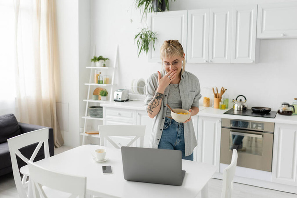 tattooed young freelancer with bangs, eyeglasses and short hair covering mouth while laughing, holding bowl with cornflakes and looking at laptop near smartphone and cup of coffee on desk in kitchen  - Photo, Image