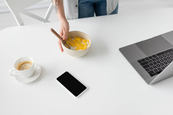 vista recortada del joven freelancer con tatuaje en el tazón de mano con copos de maíz y cuchara cerca de la computadora portátil, teléfono inteligente con pantalla en blanco y taza de café en el escritorio blanco en la cocina moderna  - Foto, imagen
