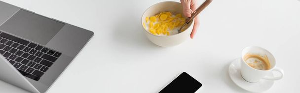 vista recortada de un joven freelancer sosteniendo un tazón con hojuelas de maíz y cuchara cerca de un teléfono inteligente portátil con pantalla en blanco y una taza de café en un escritorio blanco en la cocina moderna, pancarta  - Foto, imagen