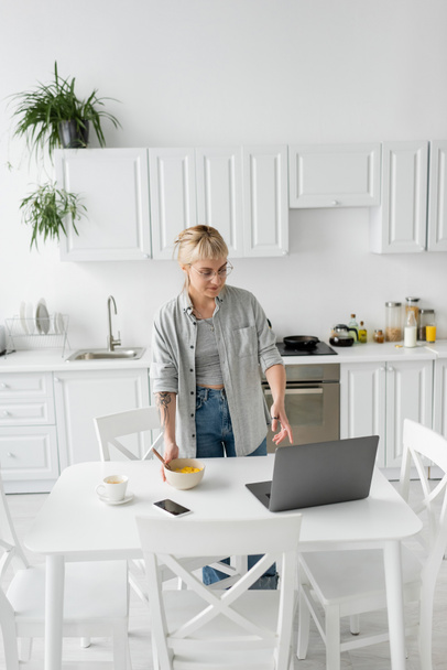 tattooed freelancer with bangs and eyeglasses holding bowl with cornflakes while having breakfast and looking at laptop near smartphone and cup of coffee on saucer and desk in modern kitchen  - Photo, Image