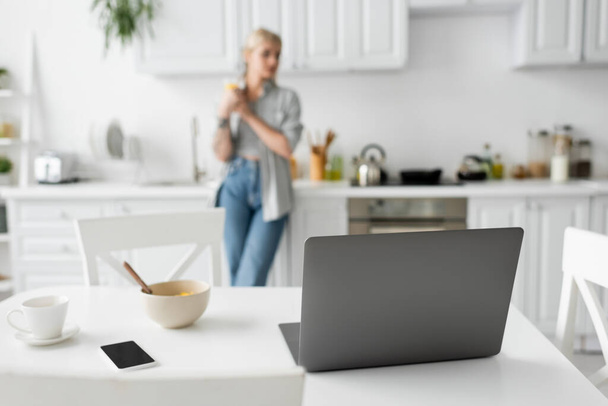 laptop, smartphone met blanco scherm, schaal met cornflakes en beker met schotel op wit bureau in de buurt van wazig freelancer staan in de moderne keuken, freelance levensstijl, digitale nomade  - Foto, afbeelding