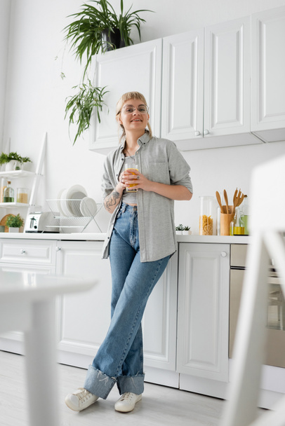 full length of tattooed and happy woman in eyeglasses holding glass of orange juice near kitchen worktop with clean dishes, toaster and rack with plants looking at camera in modern apartment  - 写真・画像