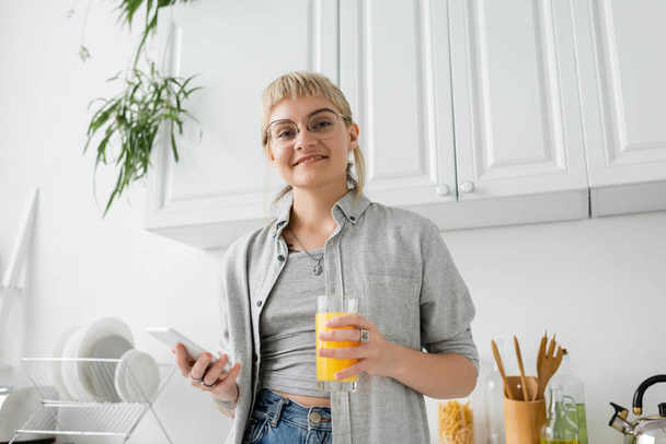 tetovaná a šťastná žena s ofinou a brýlemi držící sklenici pomerančového džusu a smartphonu při pohledu na kameru poblíž čistého nádobí a rozmazaných zelených rostlin v moderním bytě  - Fotografie, Obrázek
