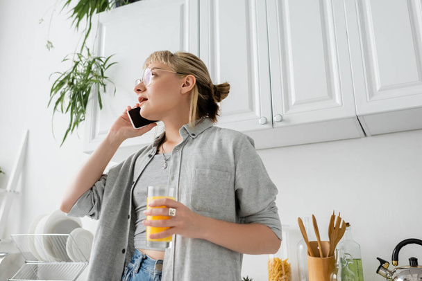 low angle view of young woman with bangs and eyeglasses holding glass of orange juice and talking on smartphone while looking away and standing in kitchen and blurred green plants in modern apartment - Photo, Image