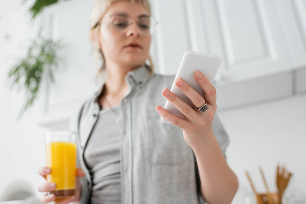 young woman in eyeglasses, with ring on finger holding glass of orange juice and smartphone while texting and standing in blurred white kitchen with green indoor plants in modern apartment  - Fotografie, Obrázek