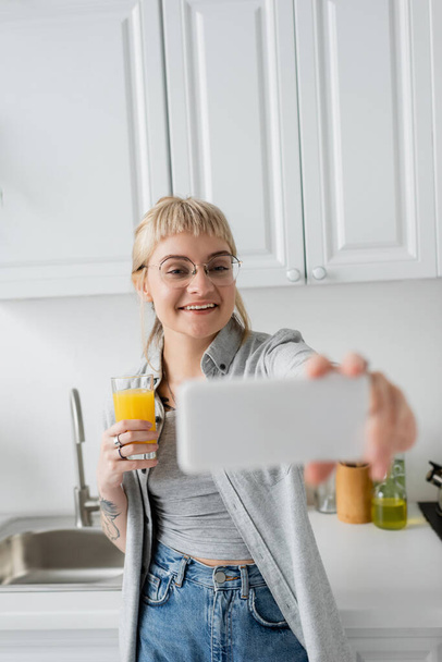 happy and tattooed young woman with bangs and eyeglasses holding glass of orange juice and taking selfie on blurred smartphone while standing in white kitchen near sink and bottle of oil - Foto, Imagem